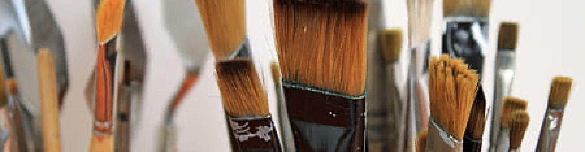 A close up shot of Canadian Artist Linda Lovisa's paint brushes, which are mostly flat and squared off style brushes on wooden handles, inside some brush holders in front of a white background.