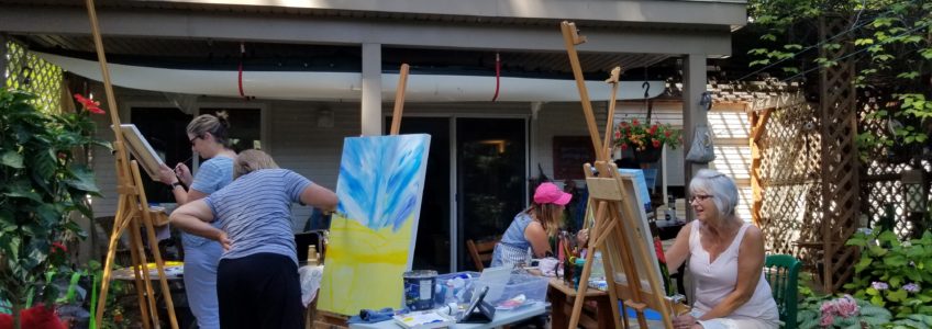 A group of people painting on canvasses outside of a house, they are at one of the classes of Linda Lovisa.