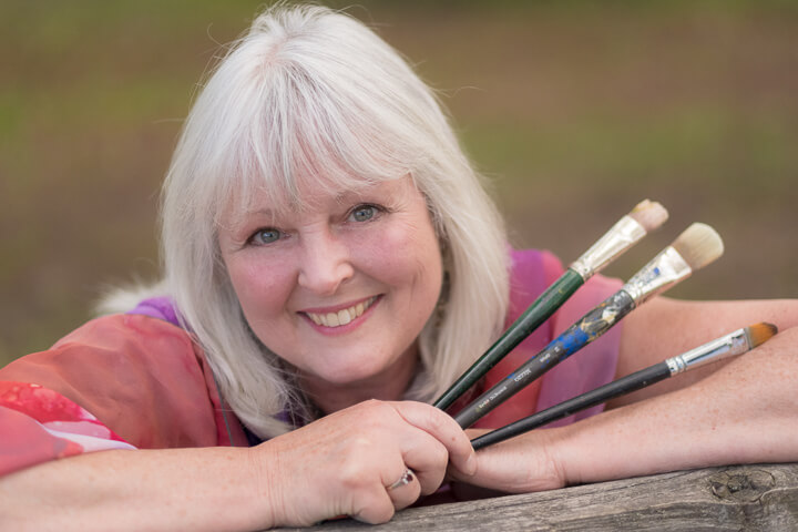 A close-up of Linda Lovisa leaning on a tree and holding three brushes.