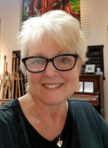 close-up portrait of Linda Lovisa, wearing black-rimmed reading glasses, a silver locket and a black shirt, and she has short white hair, with her art studio in the background.