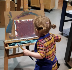 A young child wearing an apron paints scribbles across a canvas on an easel with purple paint while in Canadian artist Linda Lovisa art studio