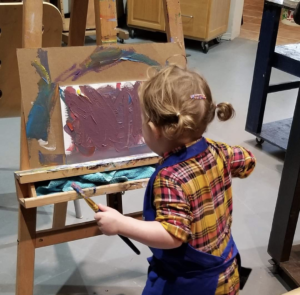 A young child wearing an apron paints scribbles across a canvas on an easel with purple paint while in Canadian artist Linda Lovisa art studio 