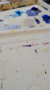 An up close look at Canadian Artist Linda Lovisa's white Matheson palette for painting with acrylics, with blue paint in small pools on it, along. with purple, yellow, red and blue stains.