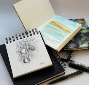 A pile of Canadian artist Linda Lovisa's sketchbooks and notebooks, with pencil drawn sketches, colour pencils sketches, scribbles and doodles, with coil bound spines and a sketch pen, on a white table.